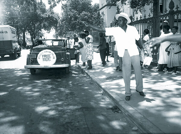 Black and White photo of a man standing in the street. Behind him is a car with the words 