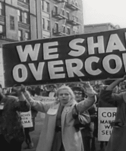 Black and white photo of a woman at a civil right's protest, holding up a WE SHALL OVERCOME sign